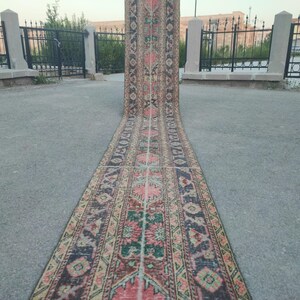 May include: A long, narrow, vintage rug with a colorful geometric pattern in shades of pink, green, black, and cream. The rug is laid out on a gray concrete surface.