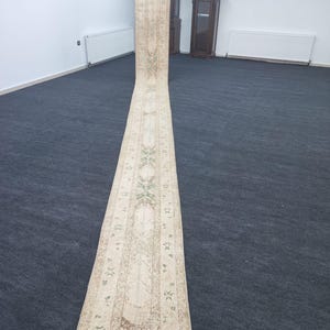 May include: A long, cream-coloured runner rug with a traditional floral pattern in shades of green and brown. The rug is rolled up and lying on a dark grey carpet.