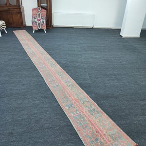 May include: A long, narrow rug with a light blue background and a pink and red geometric pattern. The rug is laid out on a dark gray carpet.