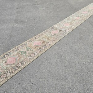 May include: A long, narrow rug with a beige background and a repeating pattern of pink, green, and brown diamonds. The rug has a distressed, vintage look.