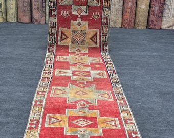 May include: A long, rectangular red rug with geometric patterns in yellow, beige, and black. The rug features a central design with diamond and triangle shapes, and a border with repeating patterns. The rug is displayed on a gray surface.