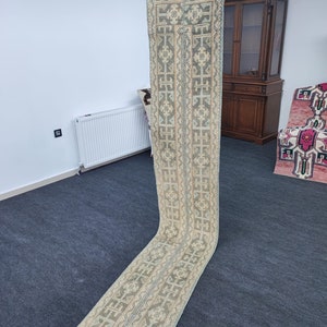 May include: A long, narrow rug with a geometric pattern in shades of beige, green, and blue. The rug is folded in half and held up by a hand.