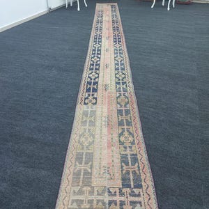 May include: A long, rectangular, vintage rug with a central pink stripe and geometric patterns in navy blue, beige, and faded green. The rug has a distressed look, with a fringed edge, and is placed on a dark gray carpet.