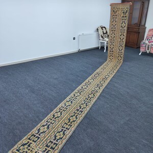 Peut inclure: Un tapis long et étroit, tissé à la main, avec un motif géométrique dans des tons de noir, jaune et orange. Le tapis est posé sur un tapis gris dans une pièce aux murs blancs.