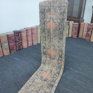 May include: A long, narrow rug with a floral pattern in shades of pink, blue, and beige. The rug is rolled up and leaning against a wall.