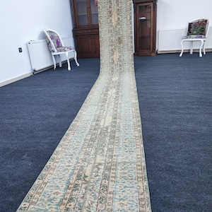 May include: A long, rectangular, vintage rug with a light blue and cream color scheme. The rug has a repeating pattern of geometric shapes and floral motifs. The rug is being held up by a person, and the bottom of the rug is laying on a dark blue carpet.