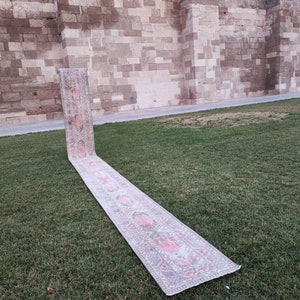 May include: A long, rectangular rug with a floral pattern in shades of pink, white, and gray. The rug is laid out on a grassy lawn in front of a stone wall.