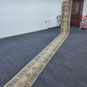 May include: A long, narrow, hand-woven rug with a traditional geometric pattern in shades of beige, brown, pink, and blue. The rug is rolled up and laid out on a grey carpet.