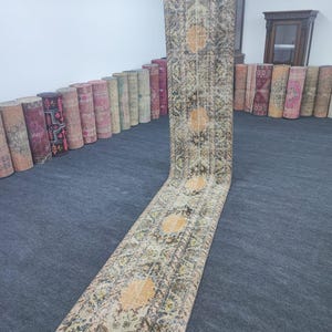 May include: A long, narrow, vintage rug with a faded floral pattern in shades of beige, brown, and blue. The rug is rolled up and standing upright on a gray surface.
