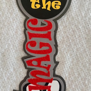 May include: A layered decoration with a black Mickey Mouse head shape at the top, the words "let the" in yellow, and the word "MAGIC" in red. The bottom features a white shape with the word "BEGIN" in blue.