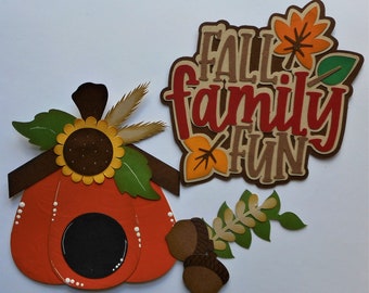 May include: Fall family fun paper craft with a pumpkin, leaves, and a sunflower. The pumpkin has a brown roof and a black circle in the center. The leaves are green and brown. The sunflower is yellow with a brown center.