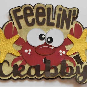 May include: A red crab with big eyes and a yellow starfish on each side. The crab is on a tan background with the words "Feelin' Crabby" in brown letters.