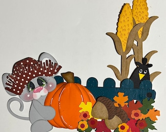 May include: A whimsical autumn-themed papercraft scene featuring a gray mouse wearing a polka-dot hat, hugging an orange pumpkin. A black bird peeks from behind a blue fence, with corn stalks, acorns, and colorful fall leaves.