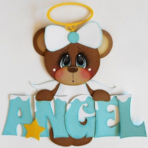 May include: A brown teddy bear with a white bow and halo, wearing a white dress and holding a blue and white sign that says "ANGEL".