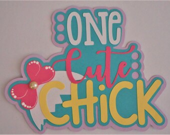May include: A pastel pink, yellow, and turquoise paper craft with the words "One Cute Chick" in white, pink, and yellow lettering. A pink flower with a pearl center is on the left side of the craft.