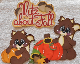 May include: A collection of fall-themed paper cutouts. The design includes two brown squirrels, pumpkins, acorns, and the phrase "Nutz about Fall" in red and orange lettering. The squirrels are holding pumpkins.