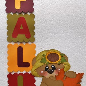 May include: A colorful fall-themed papercraft design. The word "FALL" is spelled out in individual letters with scalloped edges. A cute bear character wearing a sunflower hat holds an orange leaf. The background is white.