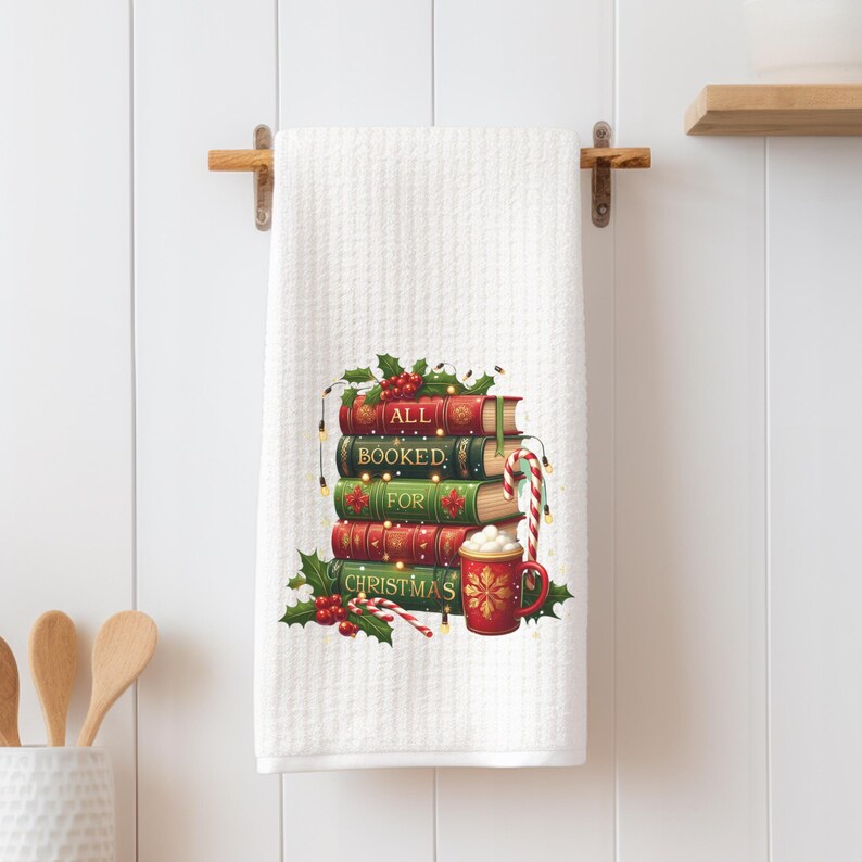 May include: White waffle-weave kitchen towel with a Christmas-themed design. The design features a stack of books with the words "ALL BOOKED FOR CHRISTMAS", holly, candy canes, and a mug of hot cocoa.