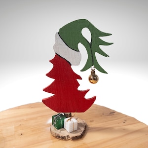 Könnte beinhalten: Eine skurrile Weihnachtsbaumdekoration. Der Baum aus Holz ist rot bemalt und hat eine grüne und weiße Mütze. Eine goldene Kugel hängt an einem grünen Zweig. Zwei kleine Geschenkboxen stehen auf einer Holzscheibe.