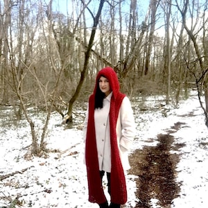 May include: A woman wearing a white coat and a long, red crocheted scarf with a matching hood. She is standing in a snowy forest.