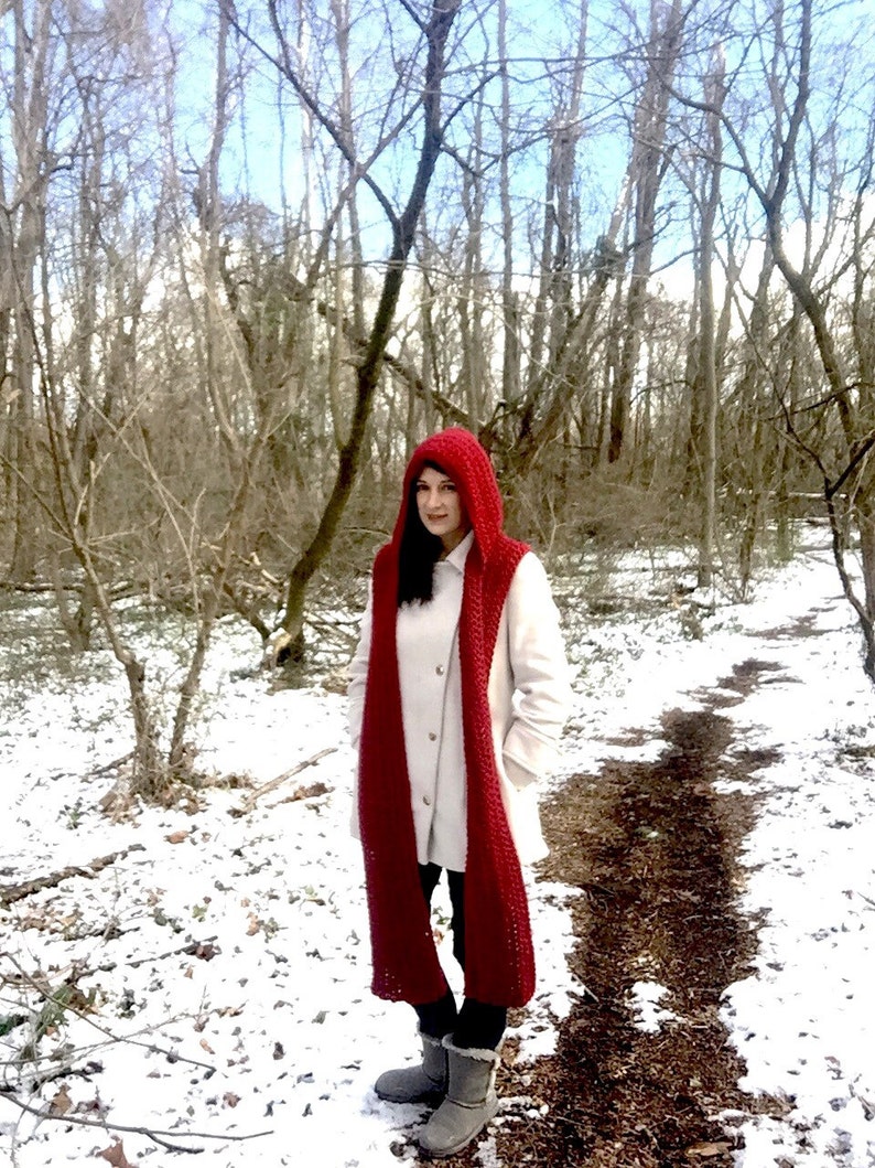 May include: A woman wearing a white coat and a long, red crocheted scarf with a matching hood. She is standing in a snowy forest.