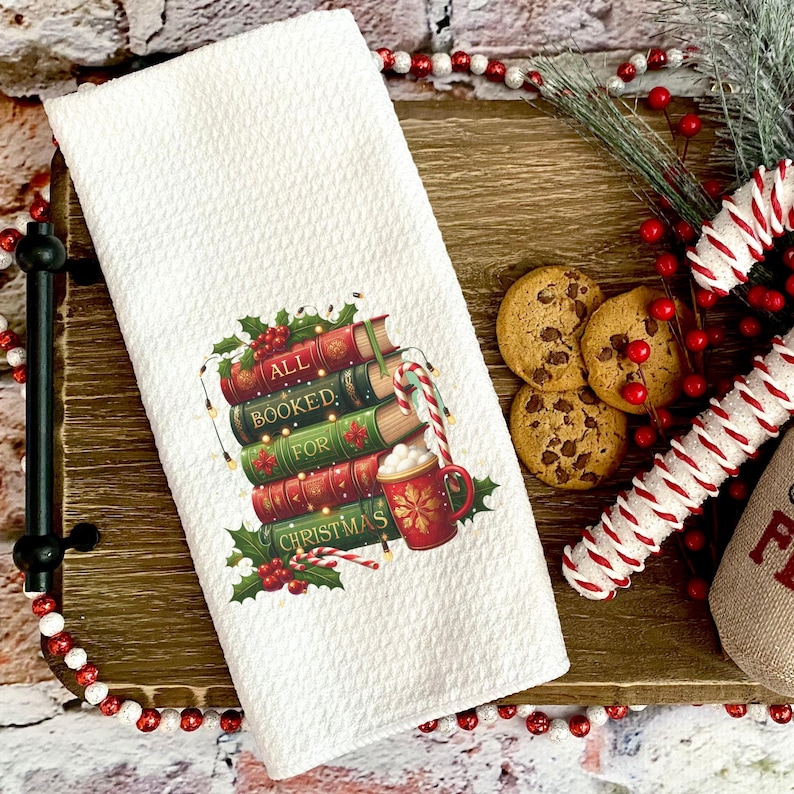 May include: White waffle-weave kitchen towel with a Christmas-themed design. The design features a stack of books with the words "ALL BOOKED FOR CHRISTMAS", a candy cane, and a mug of hot cocoa. Chocolate chip cookies and a candy cane are also visible.