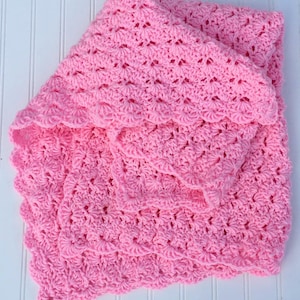 May include: A soft, bright pink baby blanket featuring a repeating shell stitch crochet pattern. The textile has a textured, scalloped edge and is folded over itself, showcasing the intricate, openwork yarn construction throughout the piece.
