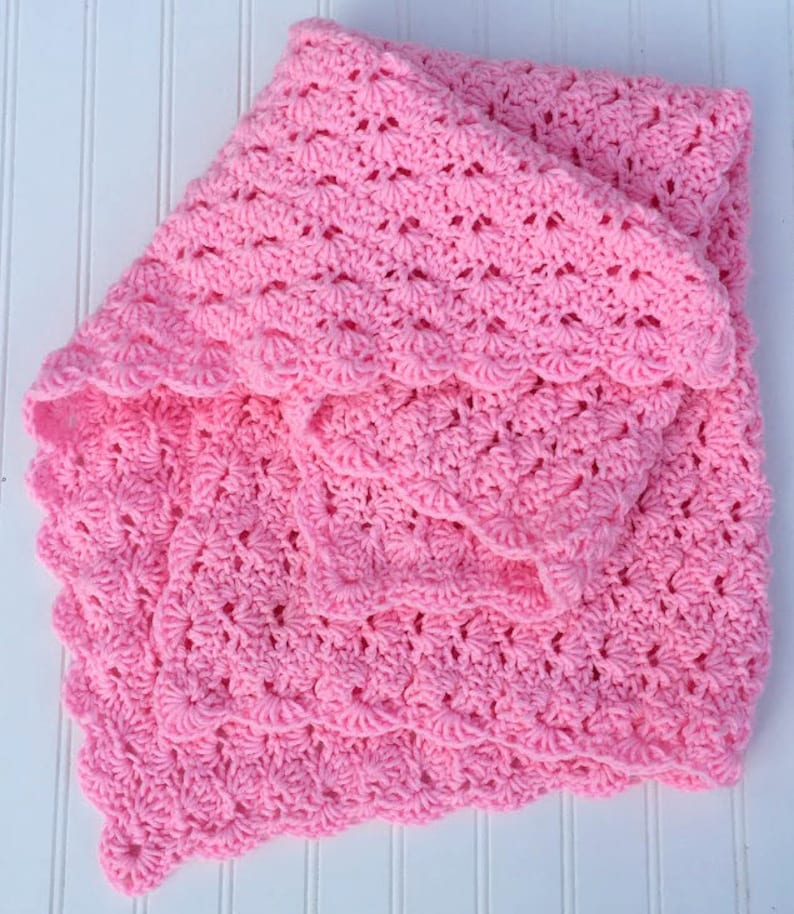 May include: A soft, bright pink baby blanket featuring a repeating shell stitch crochet pattern. The textile has a textured, scalloped edge and is folded over itself, showcasing the intricate, openwork yarn construction throughout the piece.