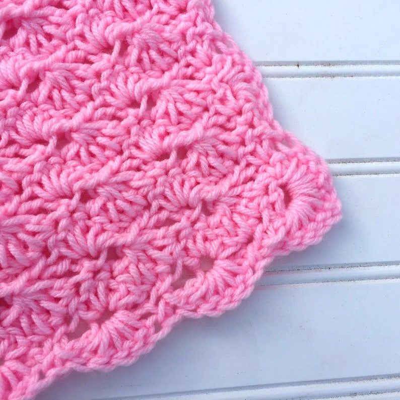 May include: A bright pink crocheted fabric featuring a repeating shell stitch pattern. The yarn is textured with visible loops and knots, creating a scalloped edge along the side of the textile piece, showcasing the intricate hand-stitched detail.