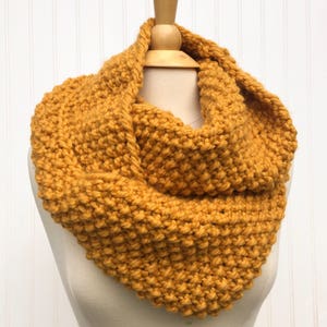 May include: A mustard yellow knitted infinity scarf. The scarf is made with a chunky yarn and has a simple stitch pattern.