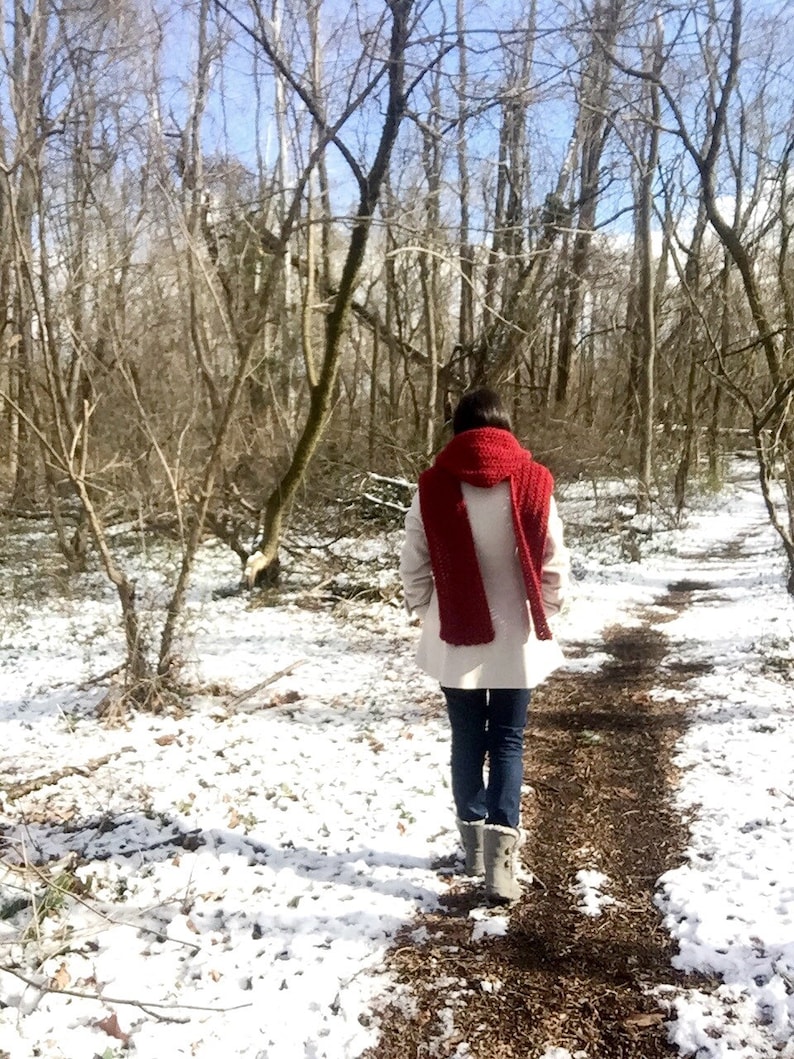 May include: A person wearing a white coat and a red scarf walks on a snowy path in a forest.