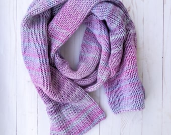 May include: A hand-knitted scarf in shades of pink, purple, and blue. The scarf is made of a thick, soft yarn with a textured appearance. The scarf is folded and draped, showcasing its length and the color variations.