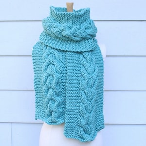 May include: A teal blue knitted scarf with a cable knit pattern. The scarf is folded over to create a cowl neck style.