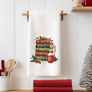 May include: White waffle-weave kitchen towel with a festive design. The design features a stack of books with the words "ALL BOOKED FOR CHRISTMAS", holly, and a mug of hot cocoa. A candy cane and string lights are also included.