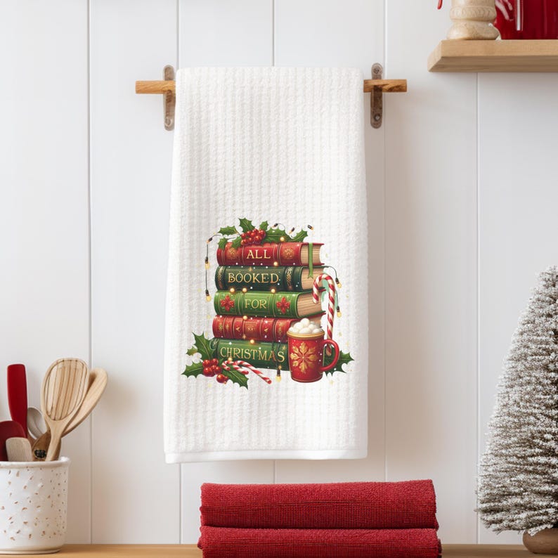 May include: White waffle-weave kitchen towel with a festive design. The design features a stack of books with the words "ALL BOOKED FOR CHRISTMAS", holly, and a mug of hot cocoa. A candy cane and string lights are also included.