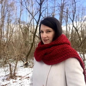 May include: A woman wearing a white coat and a long, red crocheted scarf in a snowy forest setting.