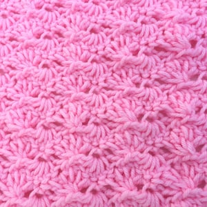 May include: A close-up view of a light pink crocheted fabric featuring a repeating shell stitch pattern. The yarn is textured and creates a series of fan-like shapes arranged in horizontal rows across the entire surface of the textile.