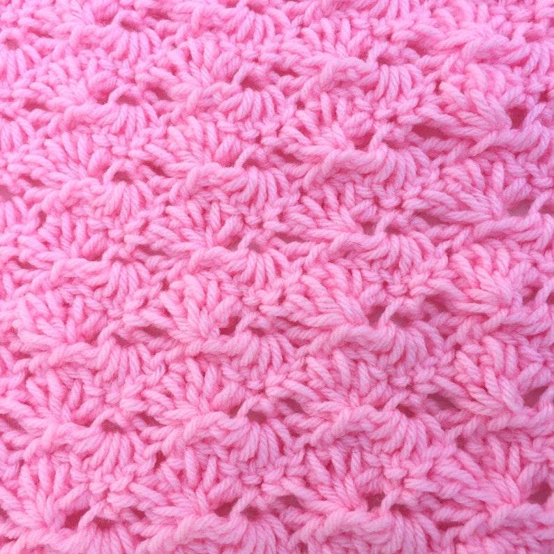 May include: A close-up view of a light pink crocheted fabric featuring a repeating shell stitch pattern. The yarn is textured and creates a series of fan-like shapes arranged in horizontal rows across the entire surface of the textile.