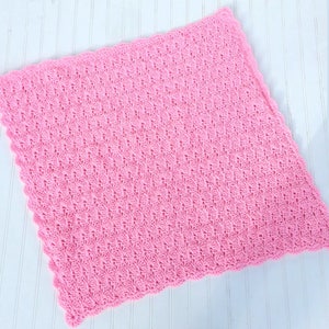 May include: A square, light pink crochet blanket featuring a repeating textured shell stitch pattern throughout the center and a scalloped edge border that frames the entire perimeter of the soft, handcrafted textile piece.
