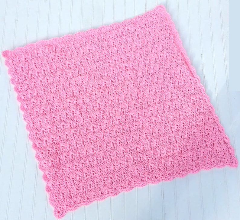 May include: A square, light pink crochet blanket featuring a repeating textured shell stitch pattern throughout the center and a scalloped edge border that frames the entire perimeter of the soft, handcrafted textile piece.