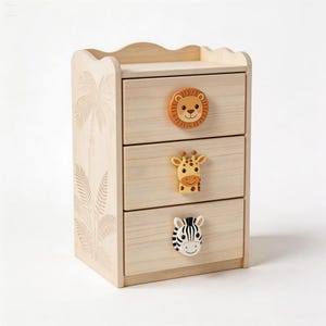 Könnte beinhalten: Eine hellfarbene Holzkommode mit drei Schubladen und Tier-Themen-Knöpfen. Die oberste Schublade hat einen Löwen, die mittlere eine Giraffe und die untere ein Zebra. Die Seite der Kommode zeigt ein geschnitztes Palmen- und Blattdesign.