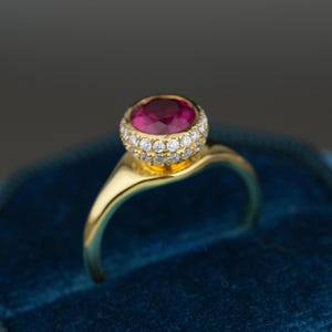以下が含まれることがあります： 大きな丸い深紅色の宝石が、小さな白い宝石のハローで囲まれた、金の指輪です。