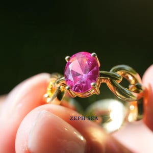 May include: A gold ring with a large, oval-cut pink gemstone. The ring has a chain-link design and the text "ZEPH SEA" is visible. The gemstone is the focal point, catching the light and reflecting a vibrant pink hue.
