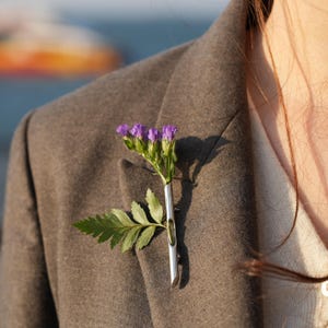 Puede incluir: Un prendedor plateado que sostiene una ramita de flores moradas y hojas verdes. El prendedor está sujeto a una blazer gris. Las flores están en plena floración y las hojas son de un verde vibrante.