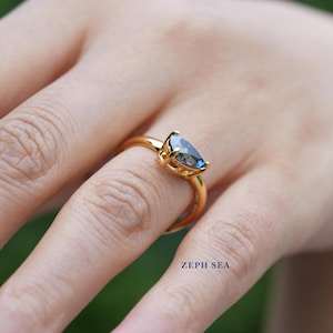 Op de afbeelding: Een gouden ring met een peervormige, blauwe edelsteen. De band en zetting van de ring zijn goudkleurig. De ring zit om een vinger. De tekst "ZEPH SEA" is zichtbaar.