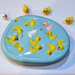 May include: A blue plastic tray filled with yellow rubber duckies, a pink rubber duck, and a white rubber duck. There are also several yellow rubber duckies on the white surface around the tray.
