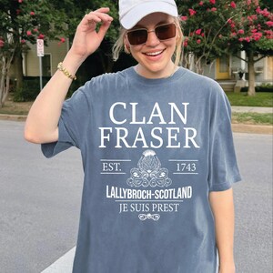 A woman wearing a white cap and sunglasses is smiling and posing in front of a house with pink flowers. She is wearing a gray t-shirt with white text and graphics printed on it.