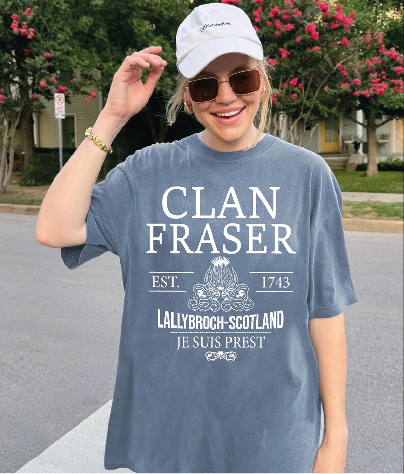 A woman wearing a white cap and sunglasses is smiling and posing in front of a house with pink flowers. She is wearing a gray t-shirt with white text and graphics printed on it.