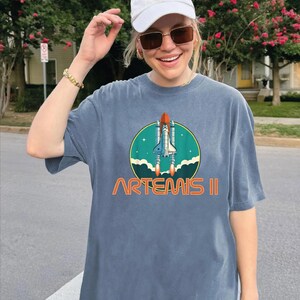 A woman wearing a white cap and sunglasses is smiling and posing in front of a house with a tree and pink flowers. She is wearing a gray t-shirt with a graphic of a space shuttle and the text "ARTEMIS II" printed on it.