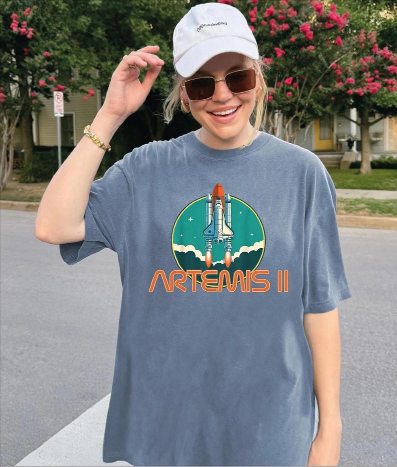 A woman wearing a white cap and sunglasses is smiling and posing in front of a house with a tree and pink flowers. She is wearing a gray t-shirt with a graphic of a space shuttle and the text "ARTEMIS II" printed on it.
